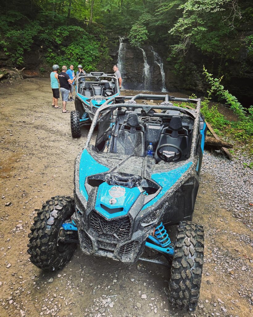 A group of people with Hatfield McCoy SXS Rentals off-road vehicles taking a break near a waterfall in a forest setting.